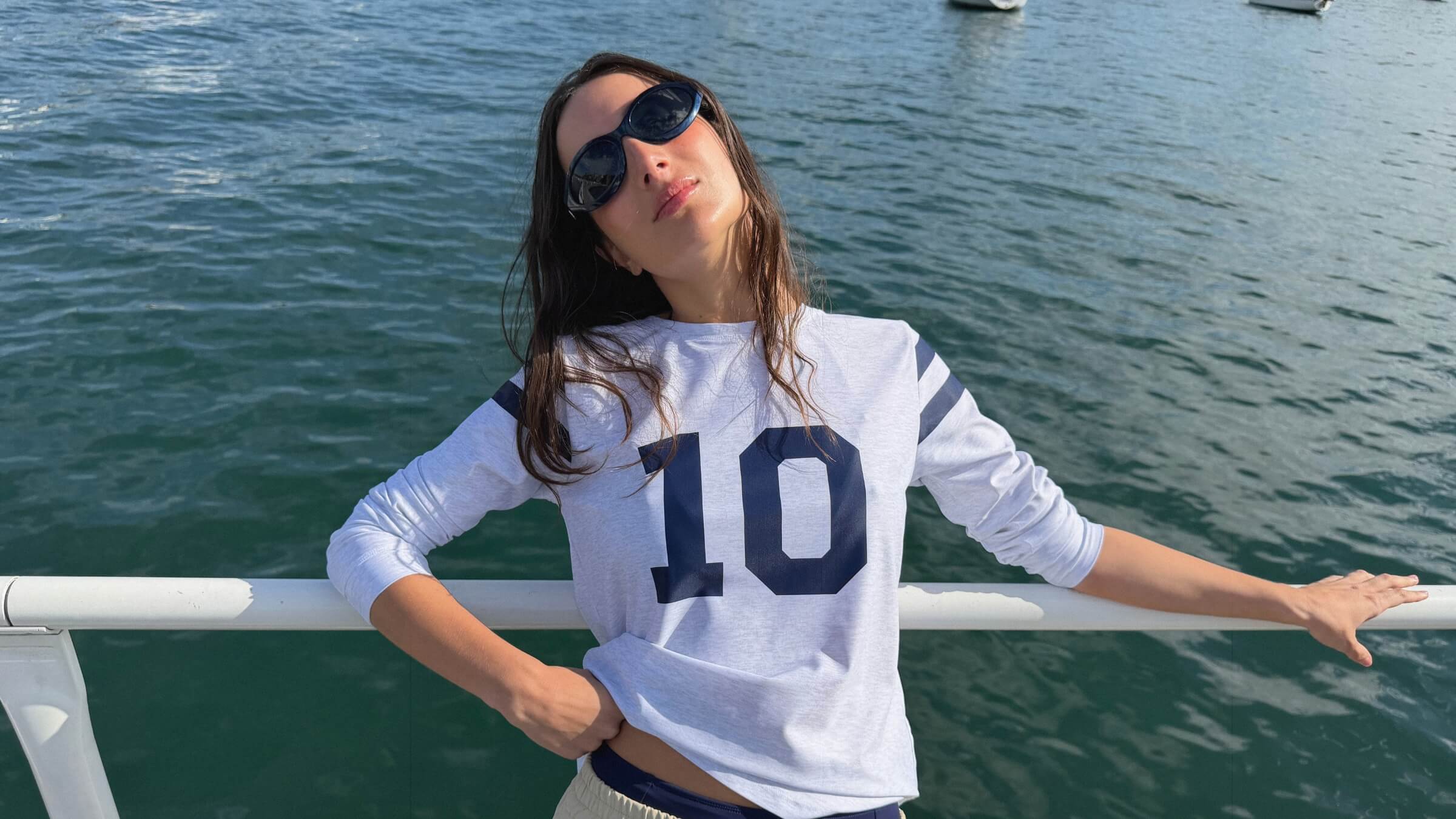 Woman wearing the ACQUA Perfect 10 UPF50+ rashguard in grey marle with navy ‘10’ print, leaning on a railing by the water.