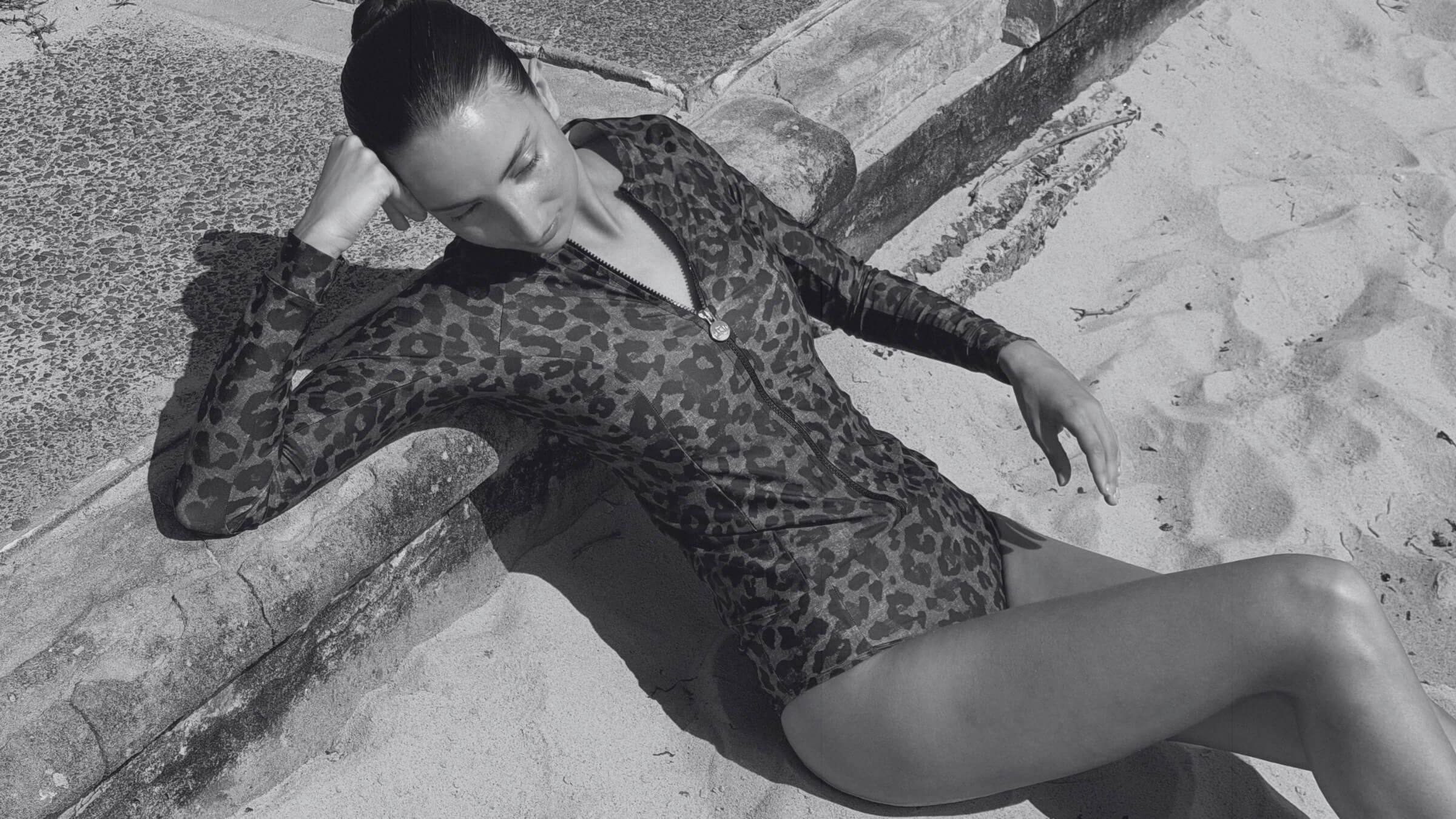 Woman wearing the ACQUA leopard print zip long sleeve UPF50+ rash guard with gold zipper, reclining on sand at the beach.