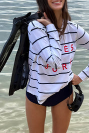 Woman wearing the ACQUA Après Surf UPF50+ rash guard in navy stripe, standing at the beach holding swim gear.