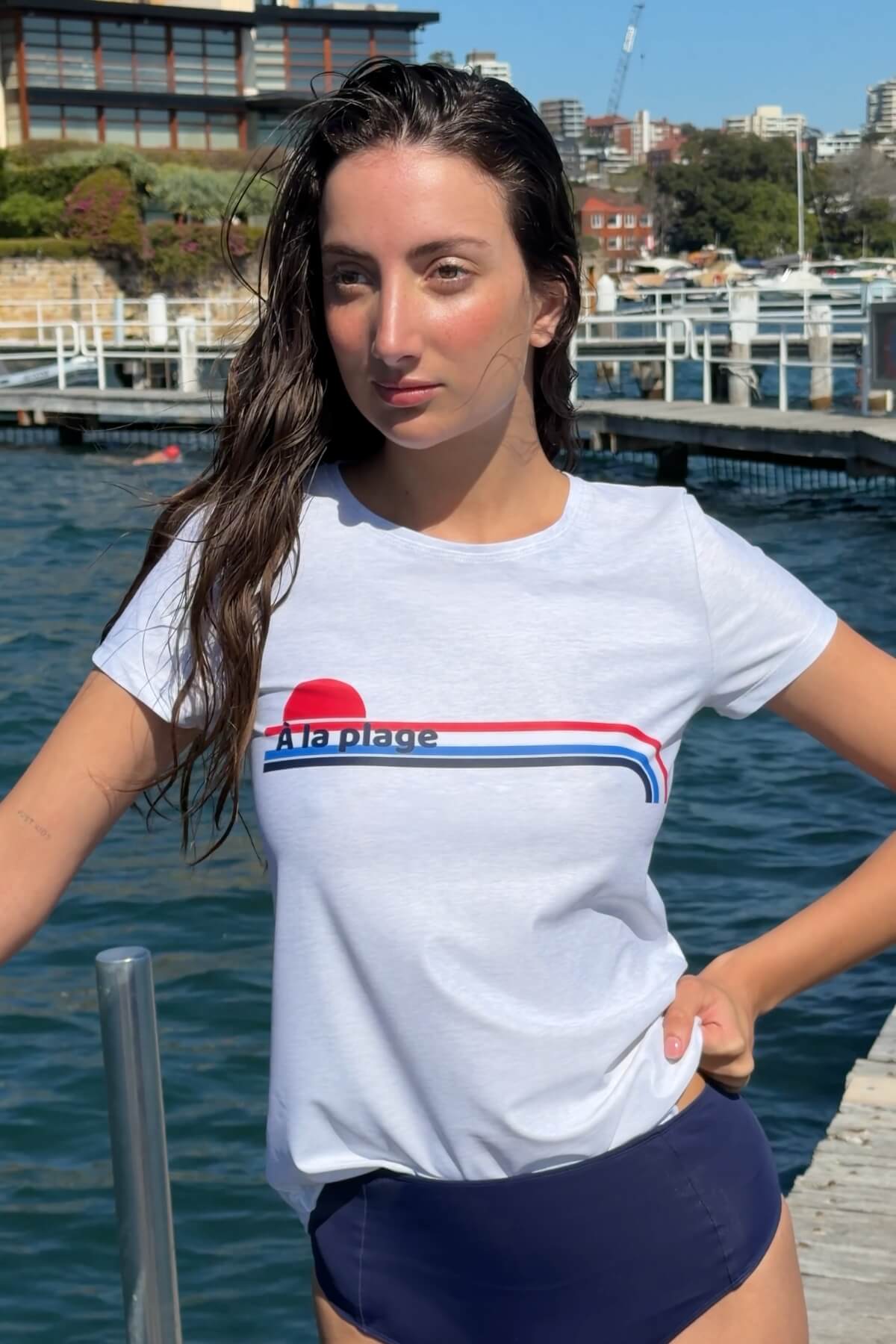 Woman wearing the ACQUA Après Surf UPF50+ rash guard in white with stripe detail, standing on a wharf by the water.