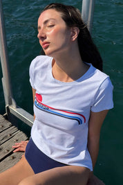 Woman wearing the ACQUA Après Surf UPF50+ rashguard in white with stripe detail, seated on a ladder by the water.