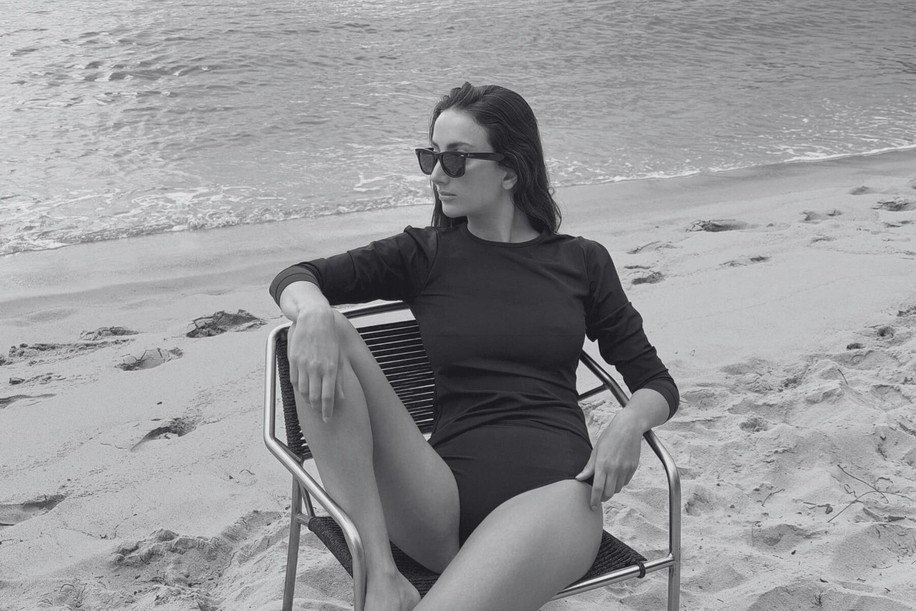 Woman wearing the ACQUA classic black UPF50+ rash guard, seated on a beach chair by the shoreline.