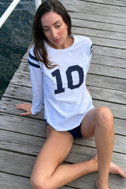 Woman wearing the ACQUA Perfect 10 UPF50+ rash guard in grey marle with navy ‘10’ print, seated on a timber jetty by the water.